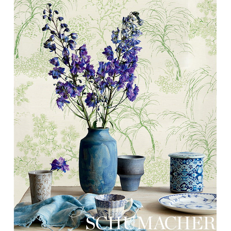 Blue ceramic vase with purple delphiniums, flanked by small blue jars, plate and cloth on a botanical wallpaper backdrop.