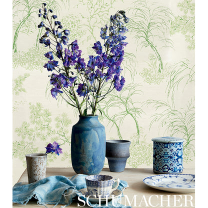 Blue ceramic vase with purple delphiniums, flanked by small blue jars, plate and cloth on a botanical wallpaper backdrop.