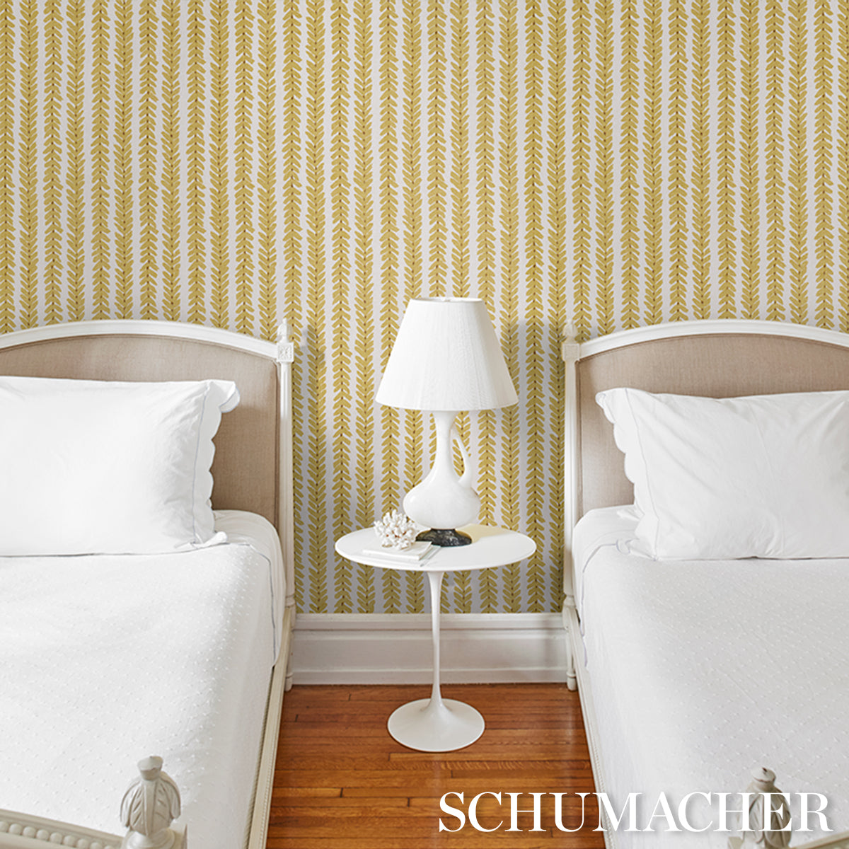 Twin bedroom with white bedding, beige headboards, gold leaf-pattern wallpaper, white round nightstand and lamp, maple floor.