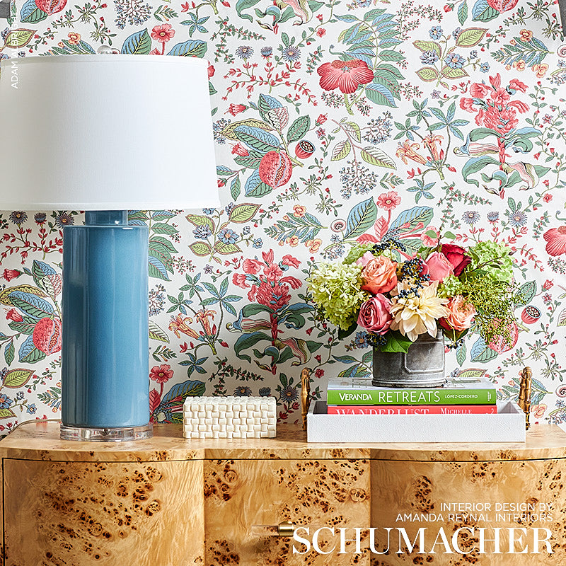 Blue ceramic table lamp with white shade beside a vase of colorful flowers on a burl wood console against floral wallpaper.