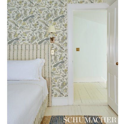 Bedroom with botanical wallpaper and birds, beige upholstered headboard, white bedding, wall lamp, light wood floor, door.