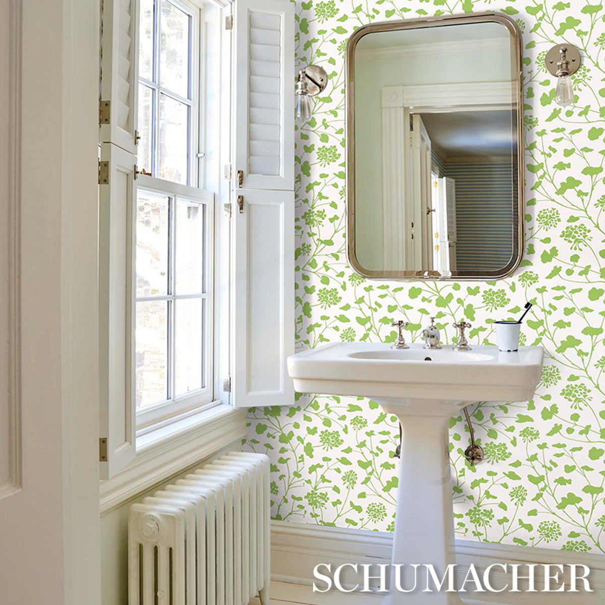 Bright bathroom with green leafy wallpaper, white pedestal sink, brass rounded mirror, and window with shutters.