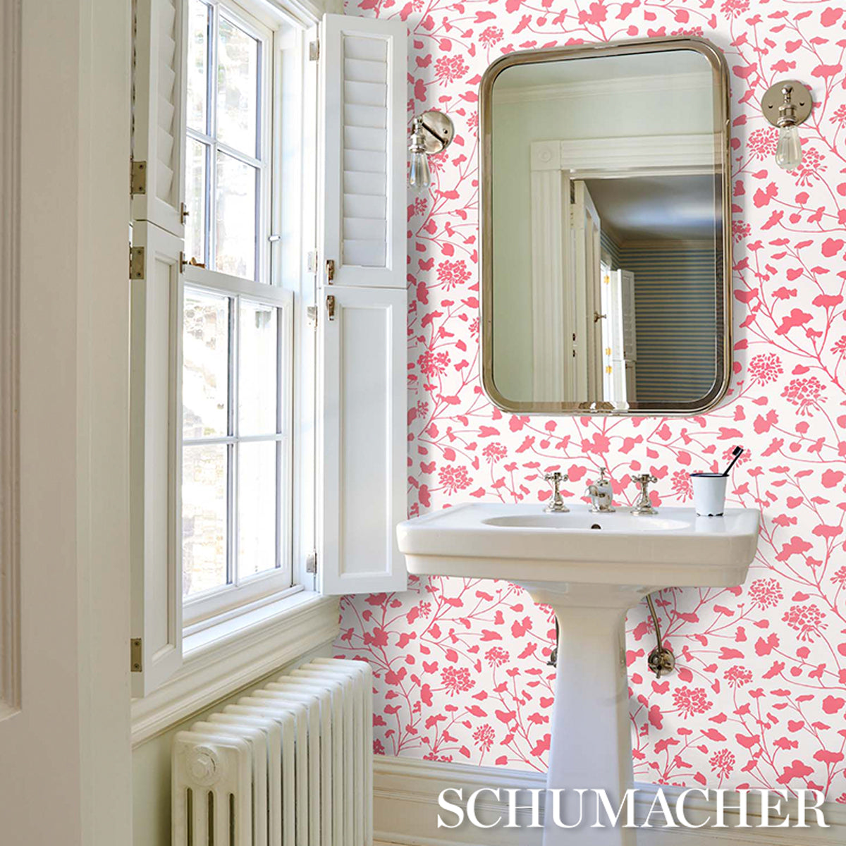 White pedestal sink with a gold-framed round mirror in a bathroom with coral-on-ivory floral wallpaper, a large window & radiator.
