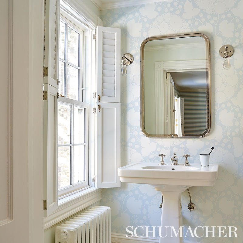 Bright vintage-style bathroom with white pedestal sink, rounded mirror, pale blue floral wallpaper, open shutters.