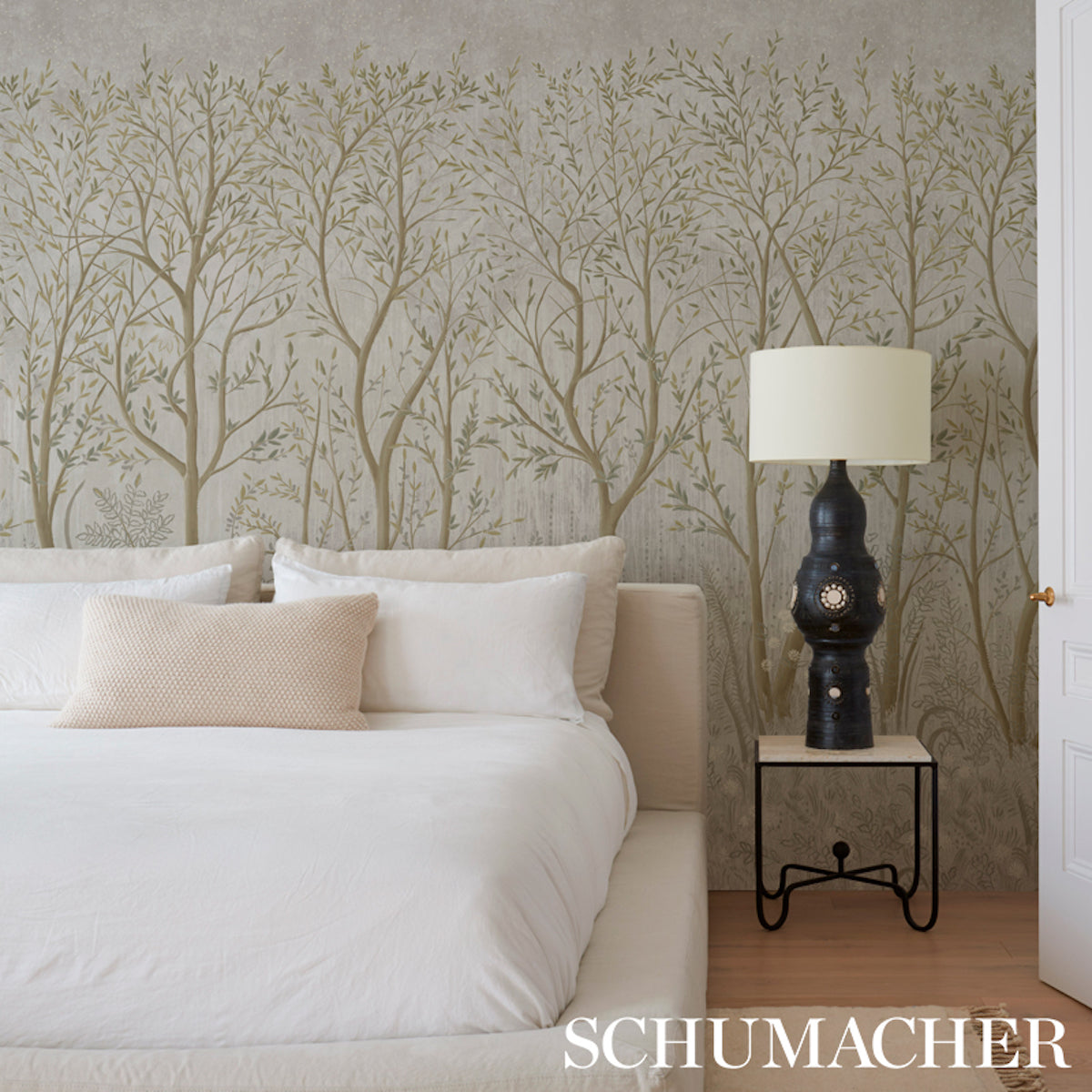 Bedroom with beige tree-pattern wallpaper, white bedding, cream pillows, and a dark ceramic lamp on a metal-frame nightstand.