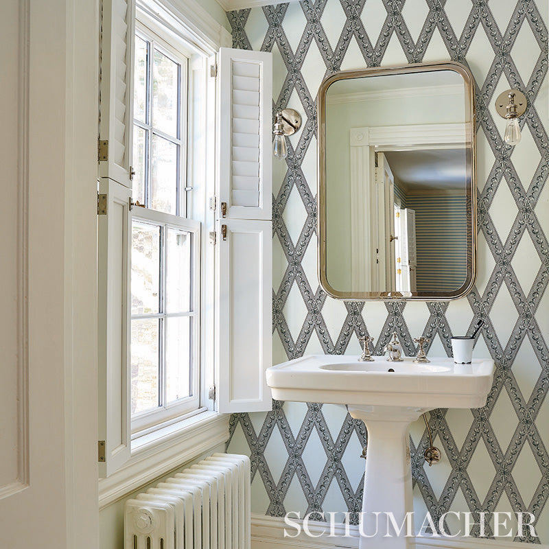 Vintage bathroom with pedestal sink, brass rounded mirror, blue-gray damask wallpaper, white shuttered window and sconces.