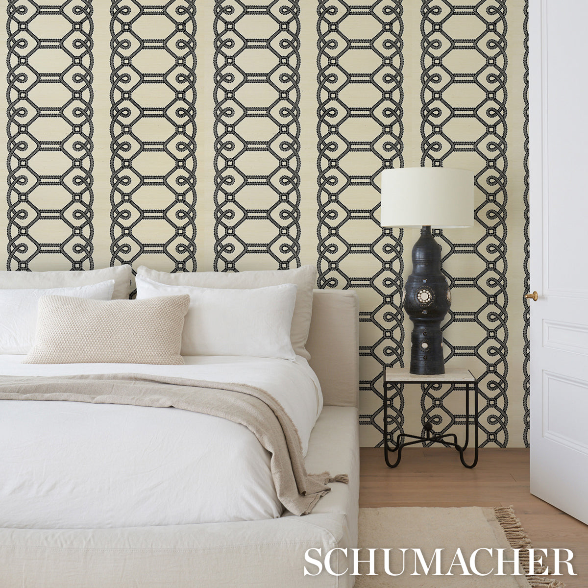 Bedroom with beige geometric chain wallpaper, white bed linens, beige throw, black lamp on metal nightstand, wooden floor.