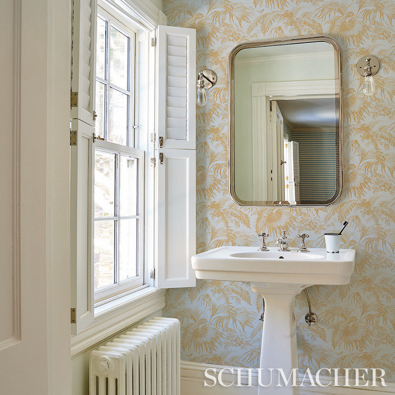 Vintage bathroom with white pedestal sink, gold-framed rounded mirror, palm-leaf wallpaper, white window with shutters.