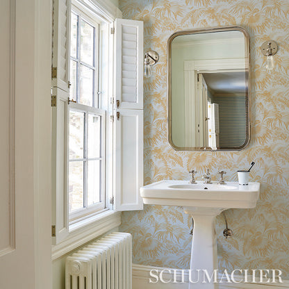 Vintage bathroom with white pedestal sink, gold-framed rounded mirror, palm-leaf wallpaper, white window with shutters.