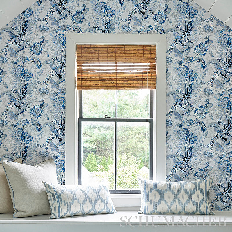 Blue-and-white floral wallpaper frames a window with a bamboo roll blind; a cozy window seat with pale blue ikat cushions.