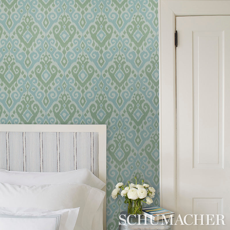 Teal-blue ikat wallpaper in a bedroom with a white upholstered headboard, white bedding, a vase of white flowers, and a door.