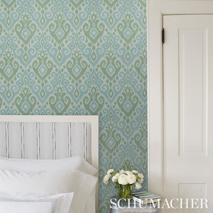 Teal-blue ikat wallpaper in a bedroom with a white upholstered headboard, white bedding, a vase of white flowers, and a door.