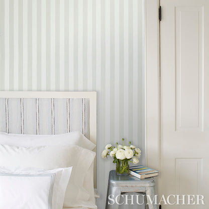 Bedroom with white bedding, striped light-gray wallpaper, metal side table, vase of white flowers and stack of books.