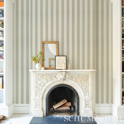 White ornate carved fireplace mantel with logs, framed art and vase on top, flanked by bookshelves and striped wallpaper.
