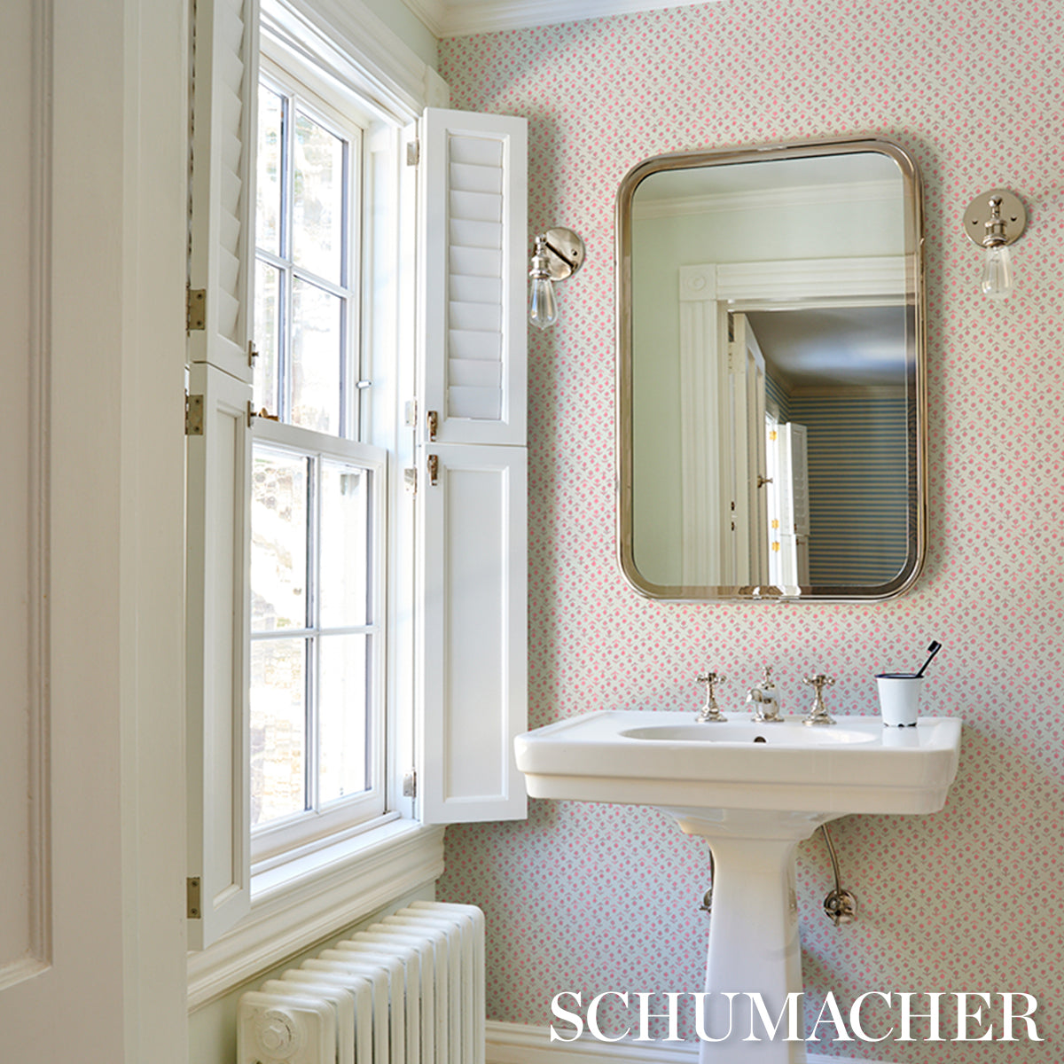 White pedestal sink with chrome taps and rounded mirror, pink dotted wallpaper, window with shutters, radiator beneath.