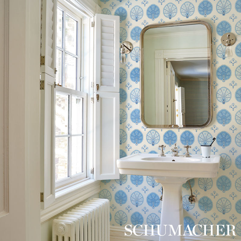 Vintage bathroom with blue wallpaper, white pedestal sink, rounded rectangular mirror, window with shutters, and radiator.