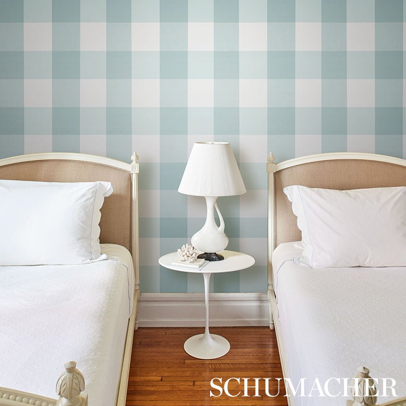 Two twin beds with beige upholstered headboards, white linens, a white nightstand and lamp between them, in a room with teal plaid wallpaper on the walls.