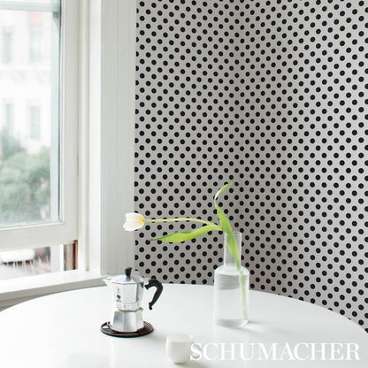 Moka pot in stainless steel on a round white table beside a glass bottle with a white tulip, against black polka-dot wallpaper.