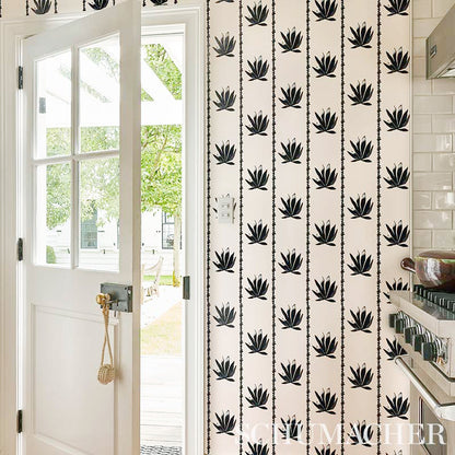 White door with glass panes opens to a green yard; beige wallpaper with black plant motifs and a white subway-tiled kitchen, rope pull.