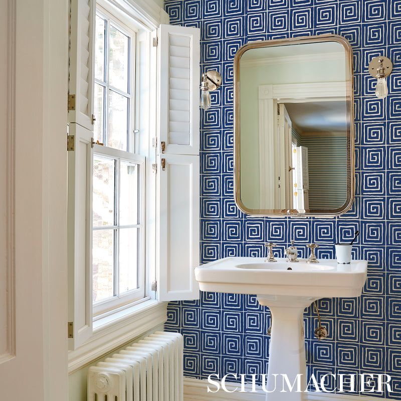 Blue Greek-key wallpaper in a vintage bathroom with a white pedestal sink, brass-framed rounded mirror, and a white shuttered window.