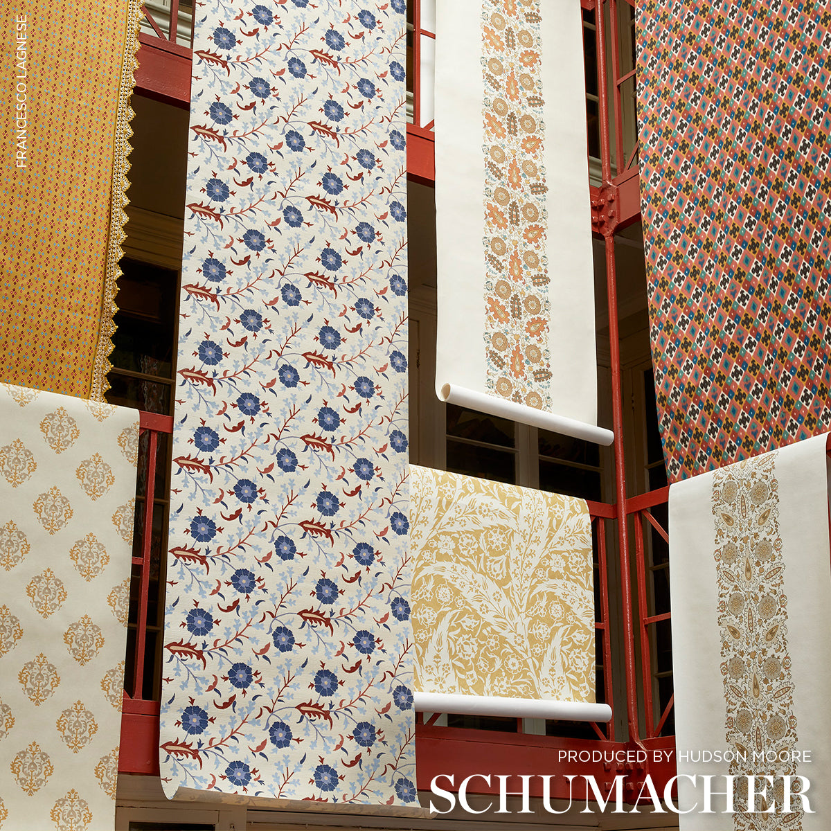 Assorted Schumacher wallpaper rolls on a red rack, featuring blue floral, ivory damask, and gold/orange patterns.