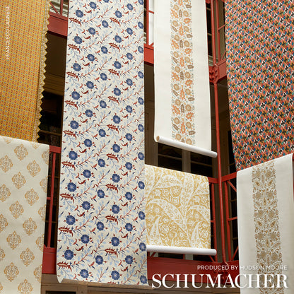 Assorted Schumacher wallpaper rolls on a red rack, featuring blue floral, ivory damask, and gold/orange patterns.