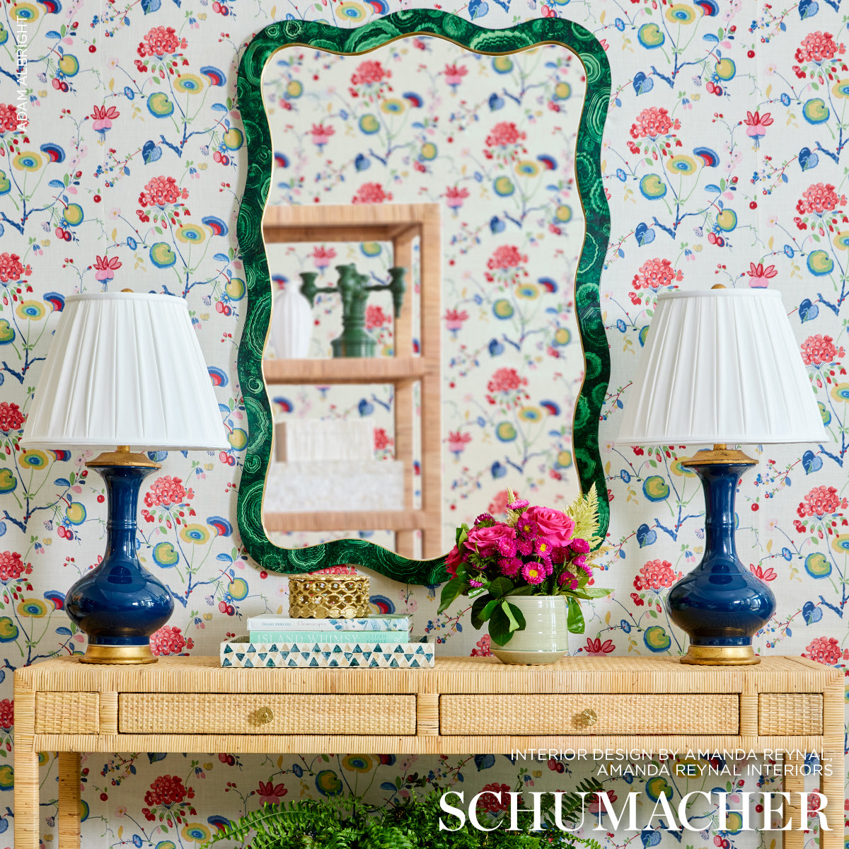 Wavy emerald-green framed mirror above a wicker console with twin cobalt lamps and pink flowers against floral wallpaper.