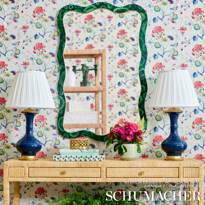 Wavy emerald-green framed mirror above a wicker console with twin cobalt lamps and pink flowers against floral wallpaper.