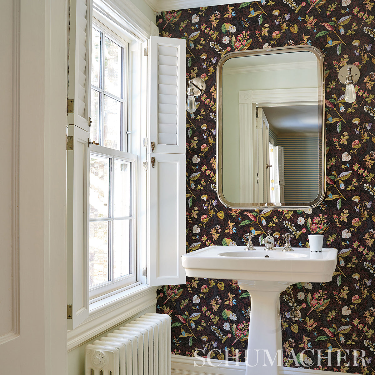 White pedestal sink with chrome taps before dark floral wallpaper and a rounded wooden-framed mirror.