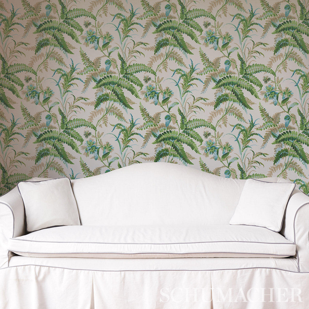 White upholstered sofa with two square cushions against tropical fern-pattern wallpaper.