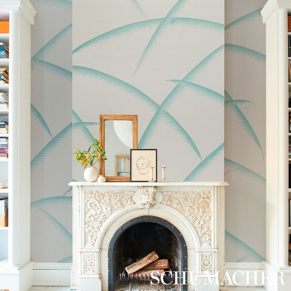 Teal palm wallpaper on a wall behind a white carved fireplace mantel; frames, a vase and candles on top, with built-in bookcases on either side.