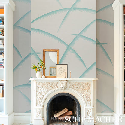 Teal palm wallpaper on a wall behind a white carved fireplace mantel; frames, a vase and candles on top, with built-in bookcases on either side.