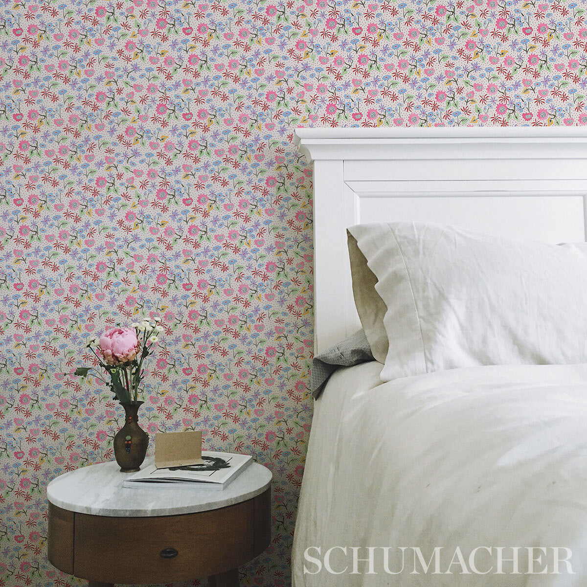 White wooden bed with a marble-topped round nightstand, pastel floral wallpaper, pink peony vase, book and card.