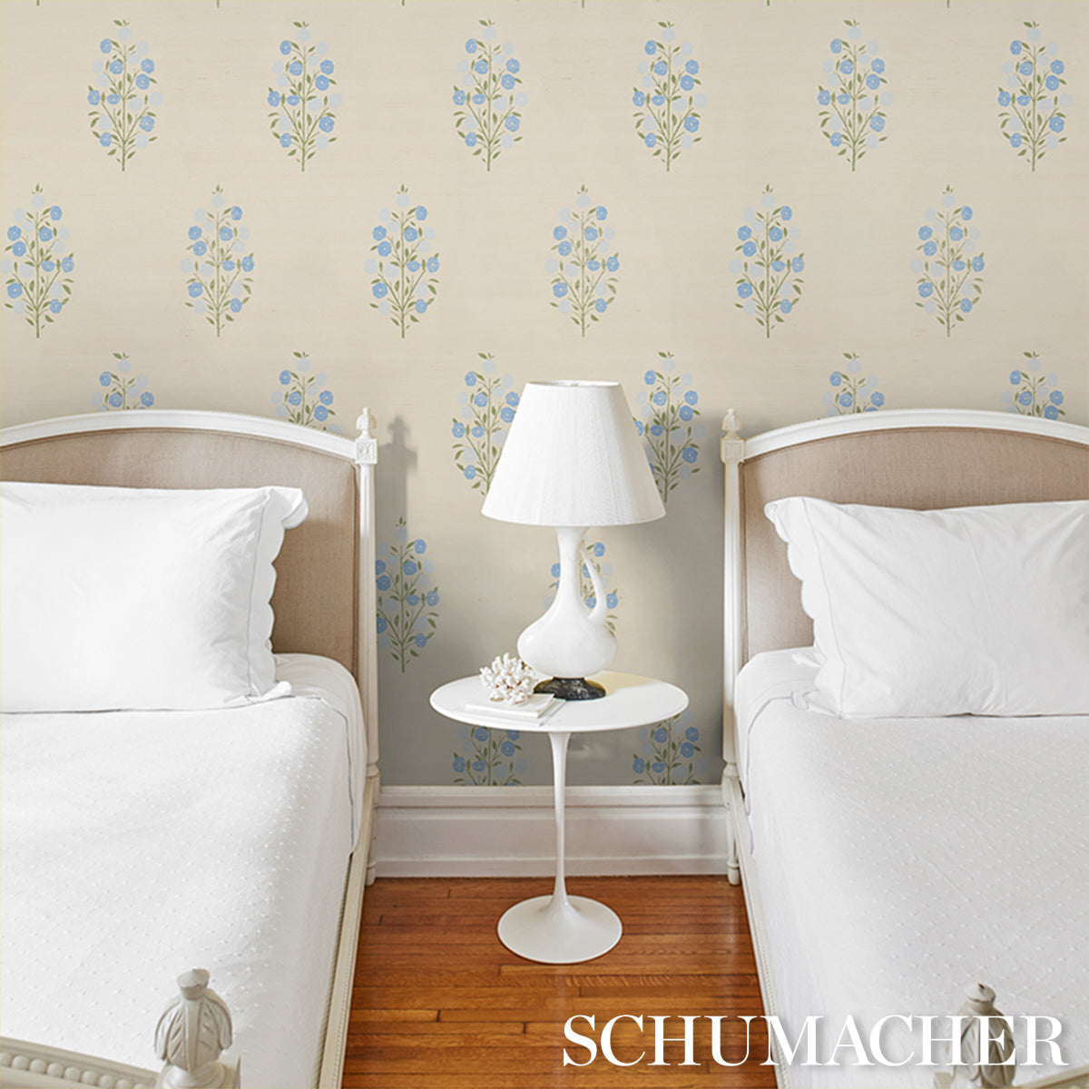 Twin bedroom with white bedding, beige upholstered headboards, white round nightstand and lamp, blue floral wallpaper.