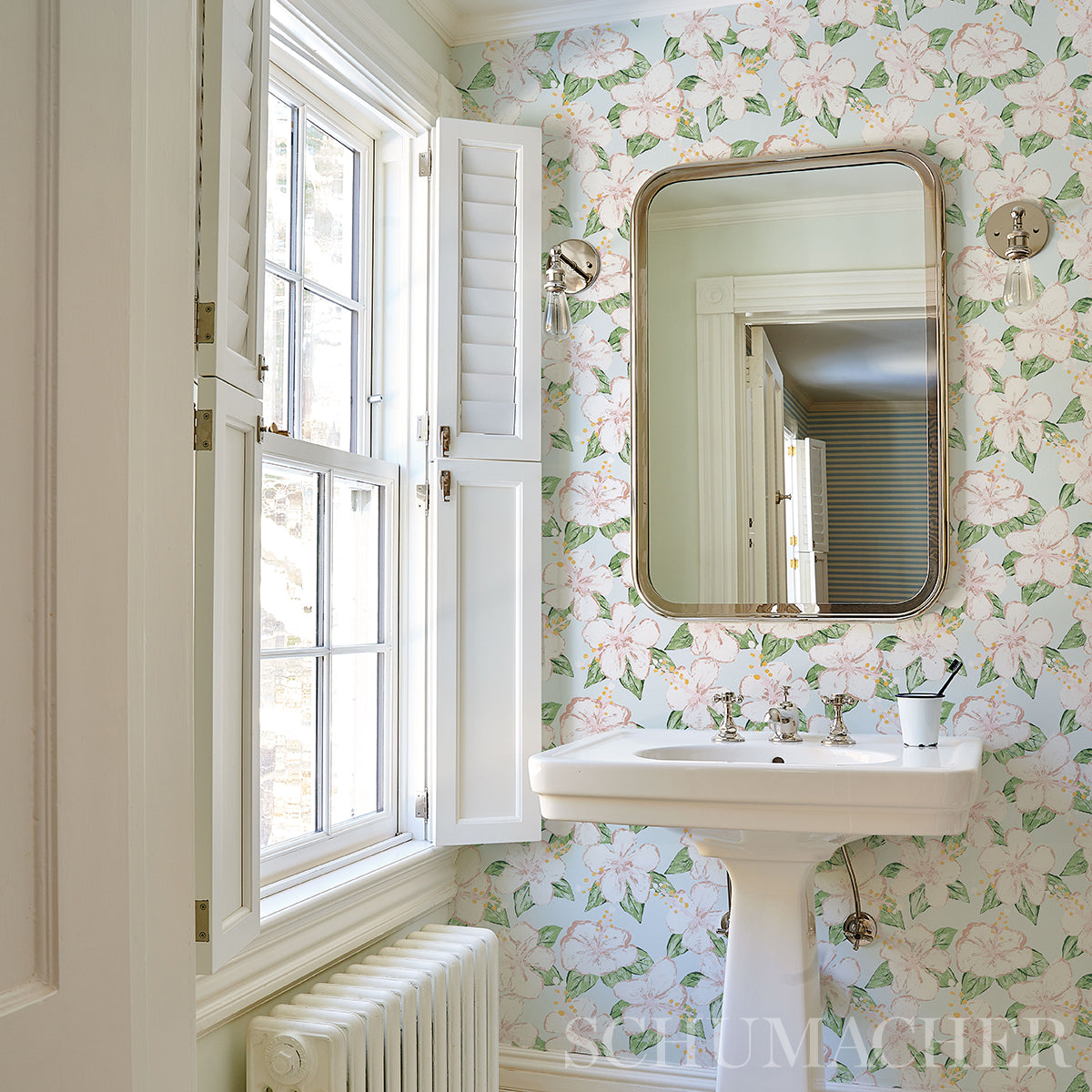 Vintage bathroom with floral wallpaper, white porcelain pedestal sink, brass-framed rounded mirror, window with shutters.