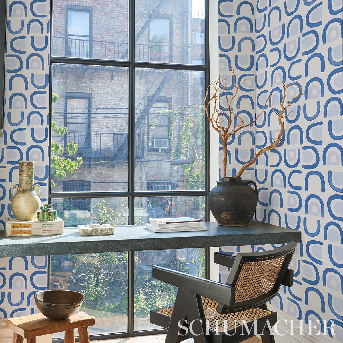 Mid-century desk by a large window, blue geometric wallpaper, grey desk, cane chair, black vase with bare branches, books.