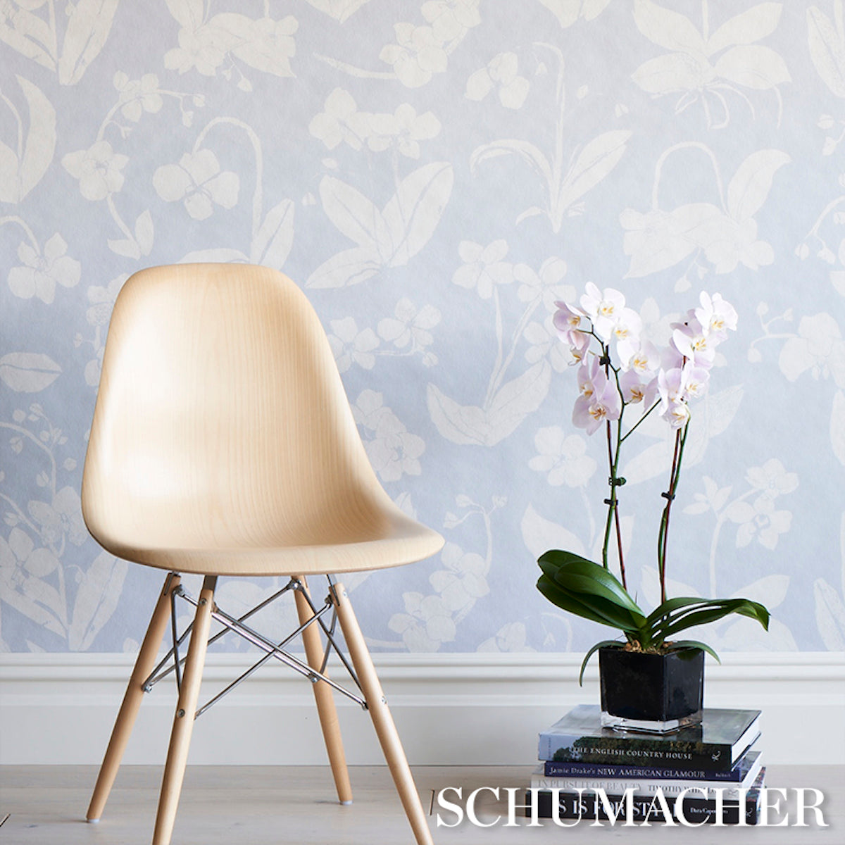 Blue orchid-pattern wallpaper with a pale sky background; in front is a natural wood mid-century chair with metal legs beside a potted orchid on books.