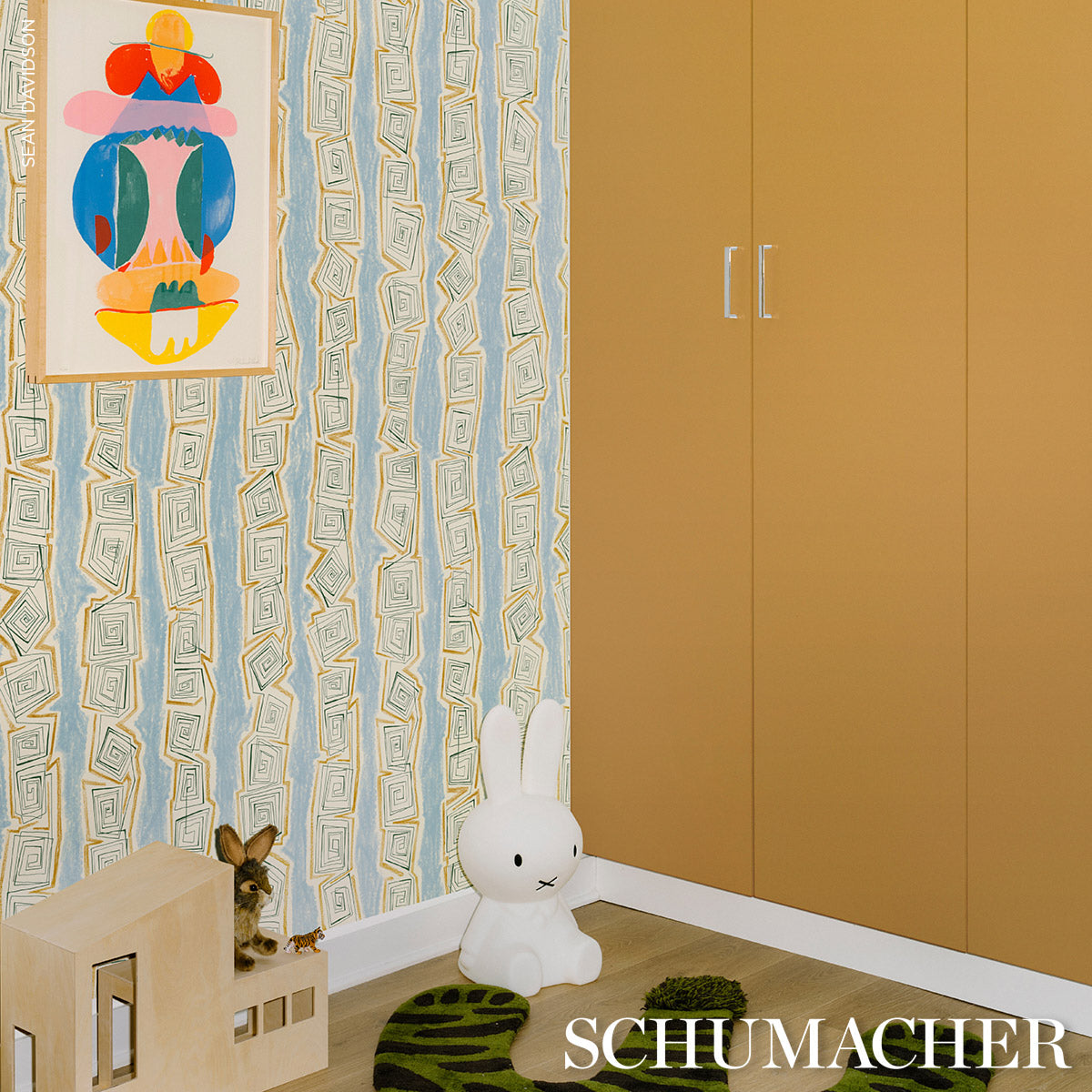 Kids' room corner with mustard wardrobe, blue geometric wallpaper, colorful art, white bunny plush, wooden playhouse.