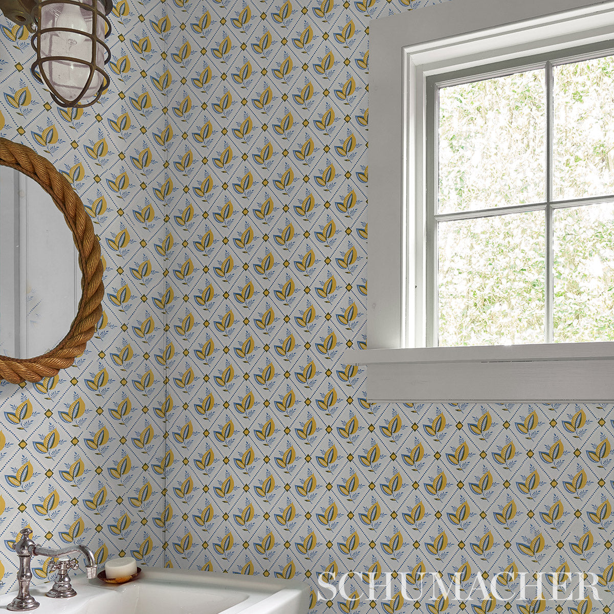 Rustic bathroom wallpaper with yellow & blue trellis pattern; rope-framed round mirror, chrome faucet sink, white window frame.