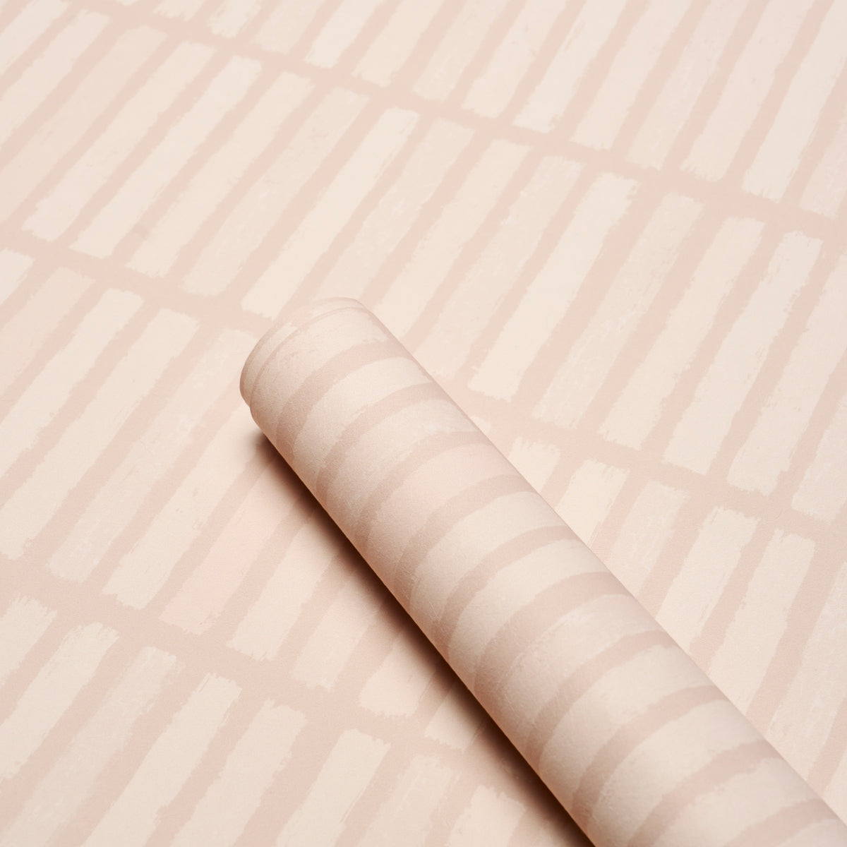 Blush pink striped wallpaper roll with diagonal bands resting on a pale pink patterned surface.
