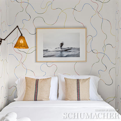 Bedroom with abstract bright multi wallpaper, amber lamp, white bedding and beige striped pillows.