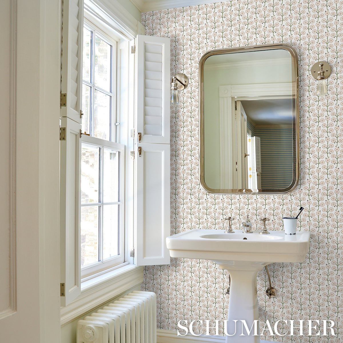 Vintage bathroom with white pedestal sink, brass-frame rounded mirror, patterned wallpaper, window shutters, and radiator.