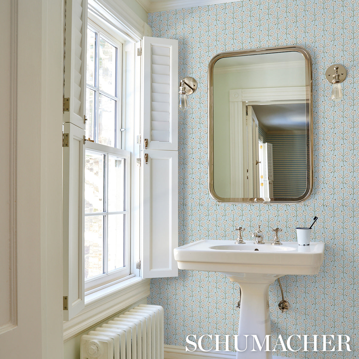 Bright vintage bathroom with pedestal sink, gold-framed rounded mirror, patterned wallpaper, sconces, window shutters.