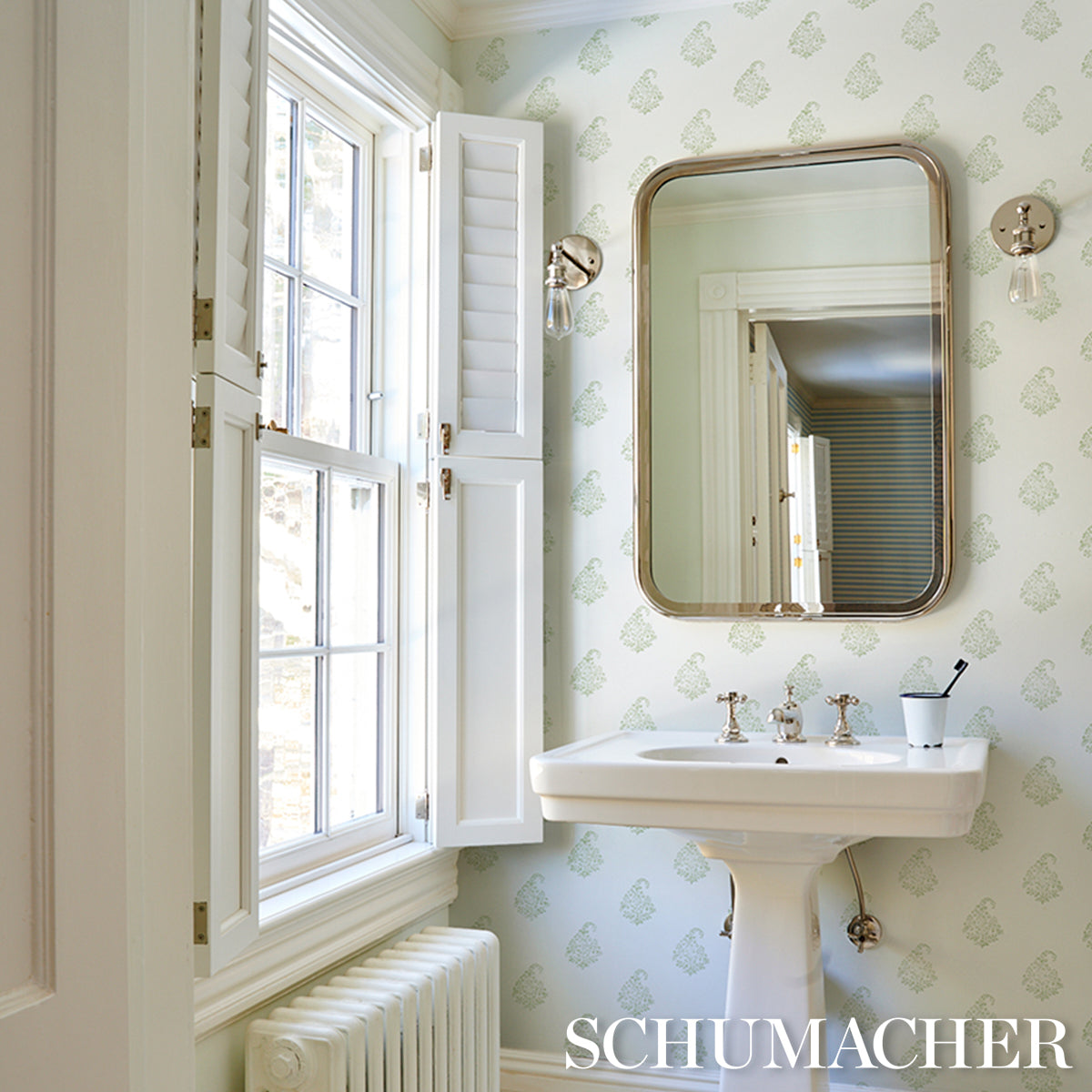 Vintage pedestal sink in a pale green damask bathroom, rounded brass-framed mirror, window with shutters, brass fixtures.