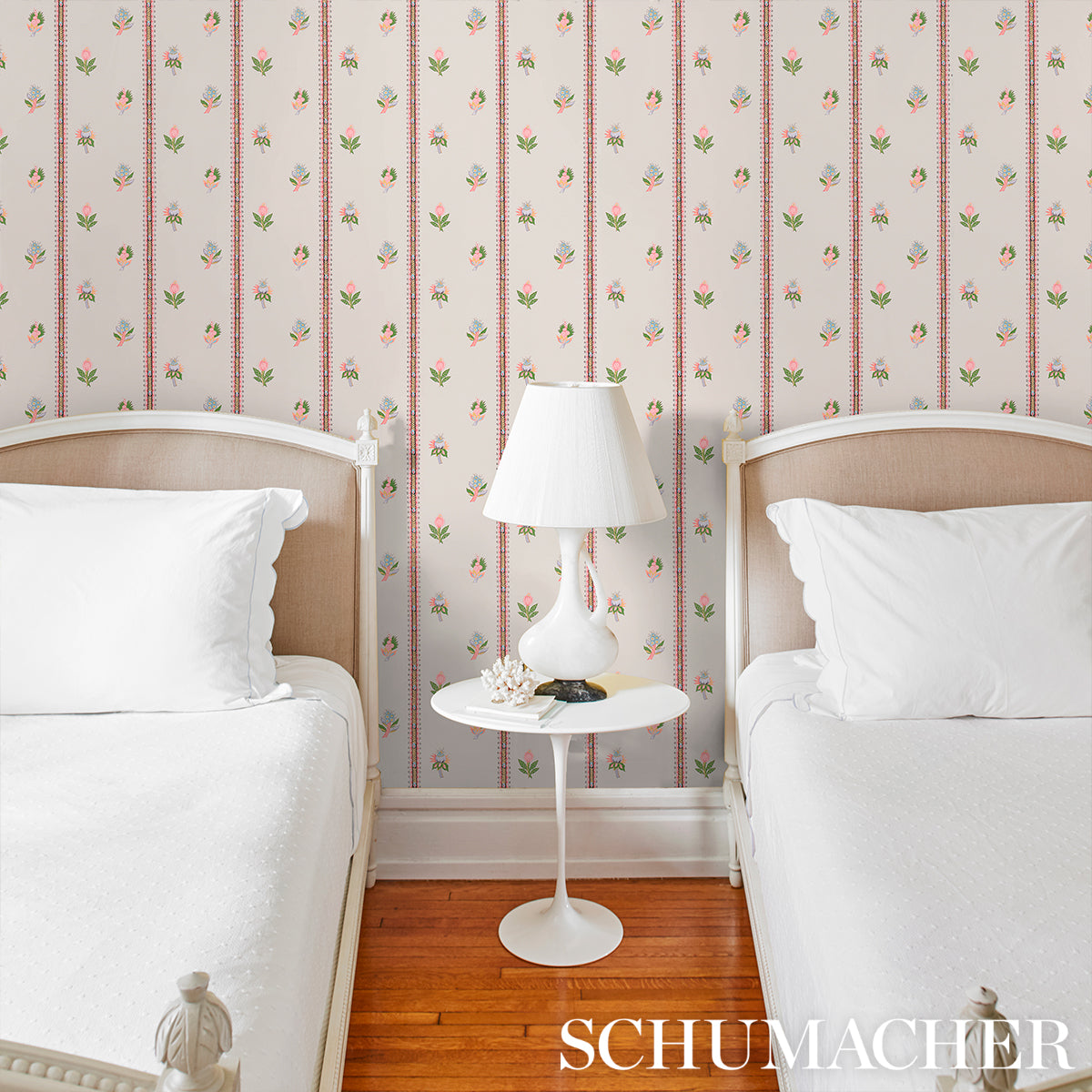 Twin beds with white bedding, white round pedestal table and lamp, pink floral striped wallpaper, wooden floor.