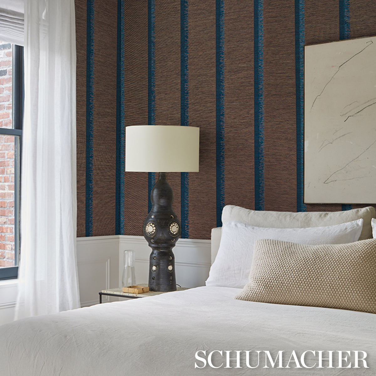 Bedroom with brown and blue striped wallpaper, white bedding, beige lumbar pillow, ornate black lamp on a nightstand.