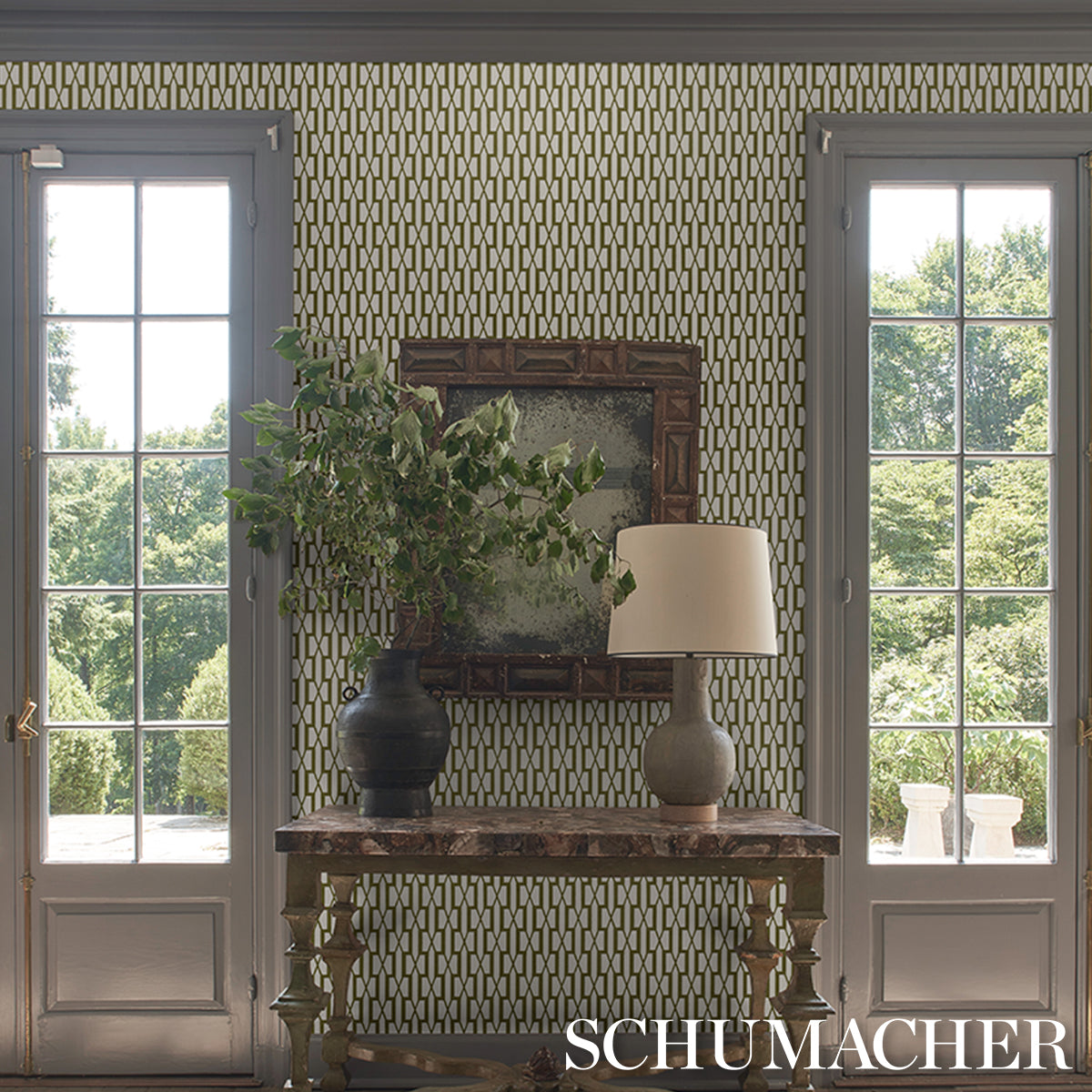 Rustic wooden console table with a greenery vase and beige lamp, ornate mirror, set against geometric Schumacher wallpaper.