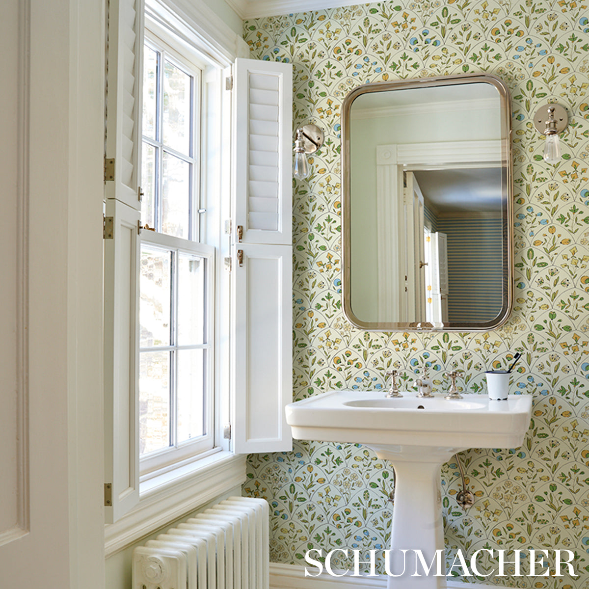 Bright retro bathroom with white pedestal sink, brass-framed rounded mirror, scallop floral wallpaper, window with shutters.