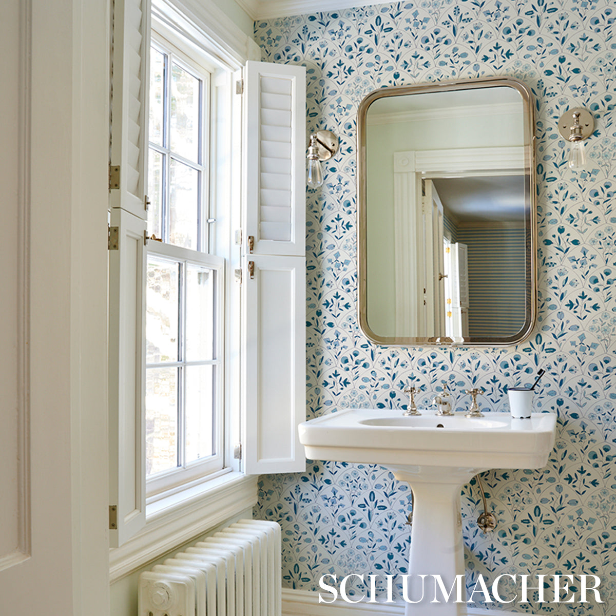 Vintage-style bathroom with white pedestal sink, brass-framed rounded mirror, blue floral wallpaper, and window with shutters.