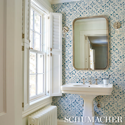 Vintage-style bathroom with white pedestal sink, brass-framed rounded mirror, blue floral wallpaper, and window with shutters.
